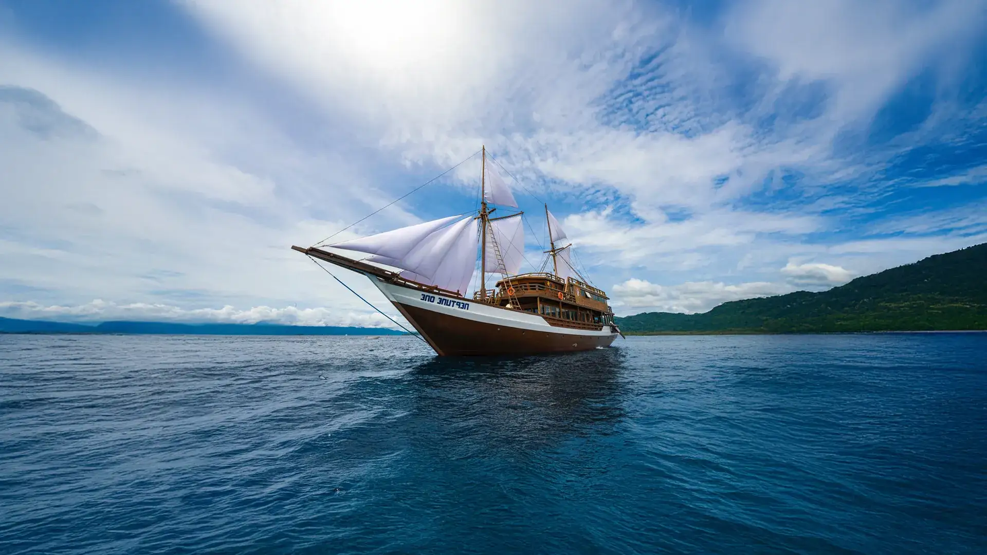 A phinisi liveaboard lying off a forested shore under open sky