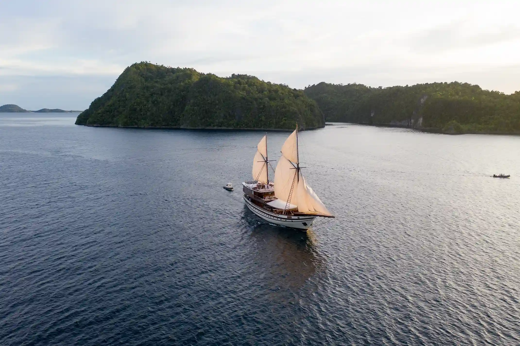 A wooden phinisi under sail beside a forested limestone island